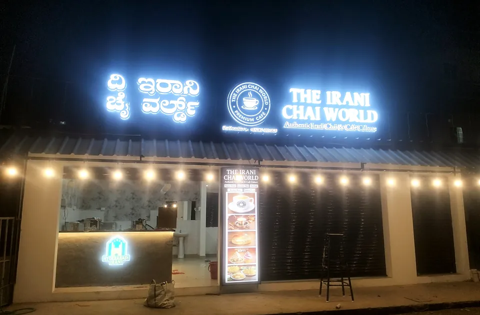 The Irani Chai World at night: illuminated 3D letters, logo and string lights on the shop front